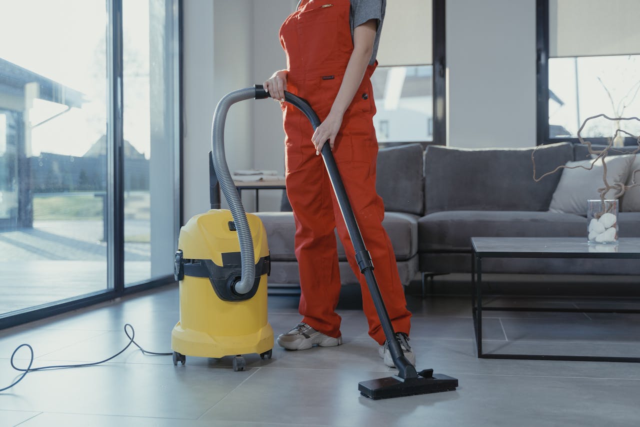 Services A professional cleaner in red coveralls vacuuming a modern living room with a yellow vacuum cleaner.