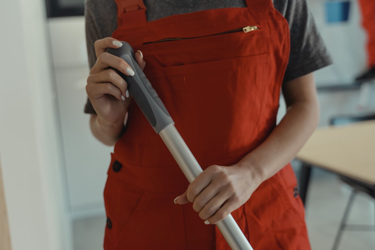 About A woman in a red apron holding a mop handle, performing cleaning indoors.